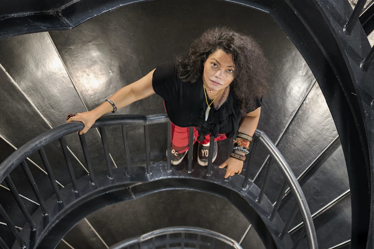 A woman stands on a metal spiral staircase, looking up frankly at the camera. She is dressed in street clothes with a bush of black hair and colourful bracelets.