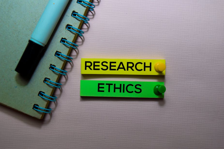 Two tags are pinned to a board next to a pen and spiral notebook. One tag, in yellow, reads "research". The other, in green, reads "ethics".