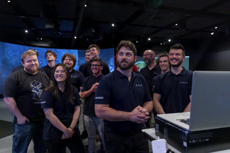A team of scientists in dark crew shirts looking at a screen above their heads