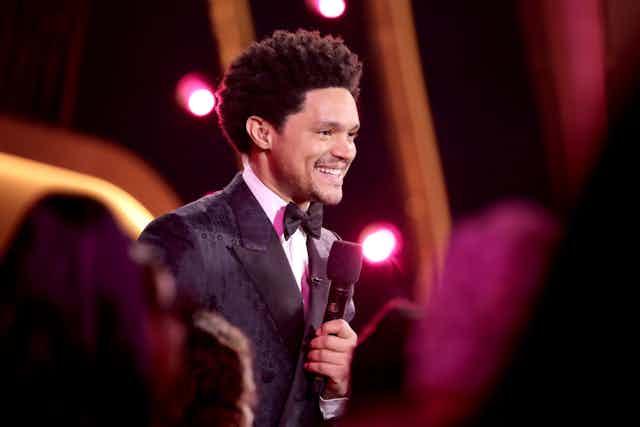 A man in a suit and bowtie with an Afro smiles as he holds a microphone, behind him stage lights.