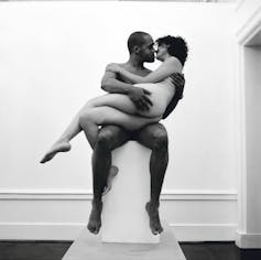 On a plinth in what appears to be an art gallery, a couple pose, about to kiss. She is 'white' and on his lap. He is 'black'. They are naked.