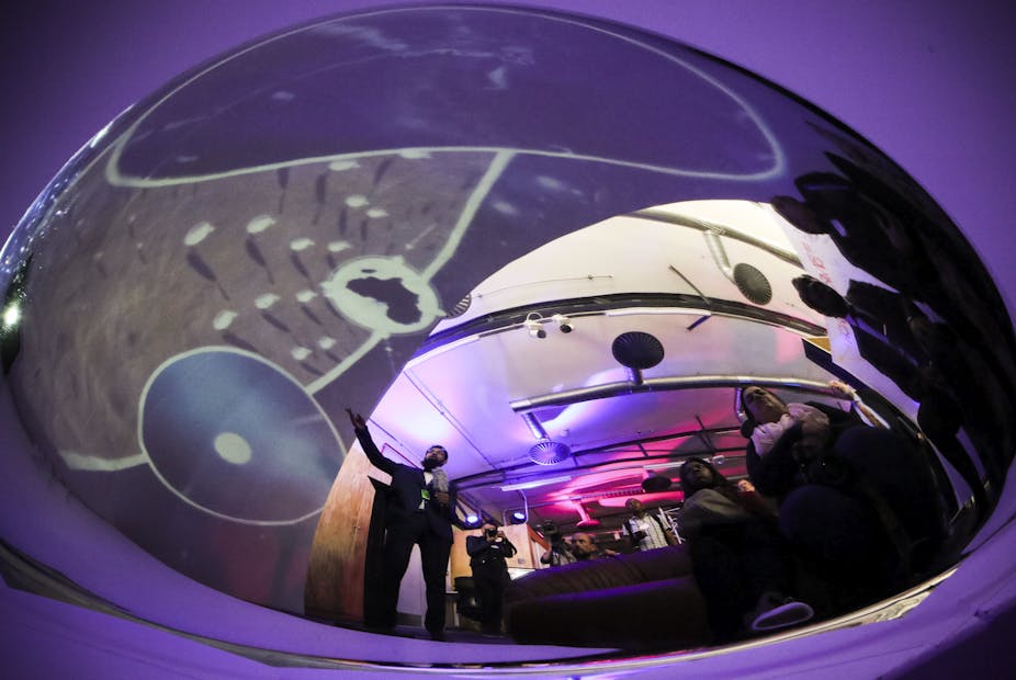 People are reflected in an item the shape of a fish bowl, which is backlit in purple