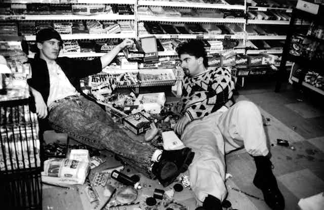 Two men sit on the floor of a convenience store surrounded by spilled merchandise.