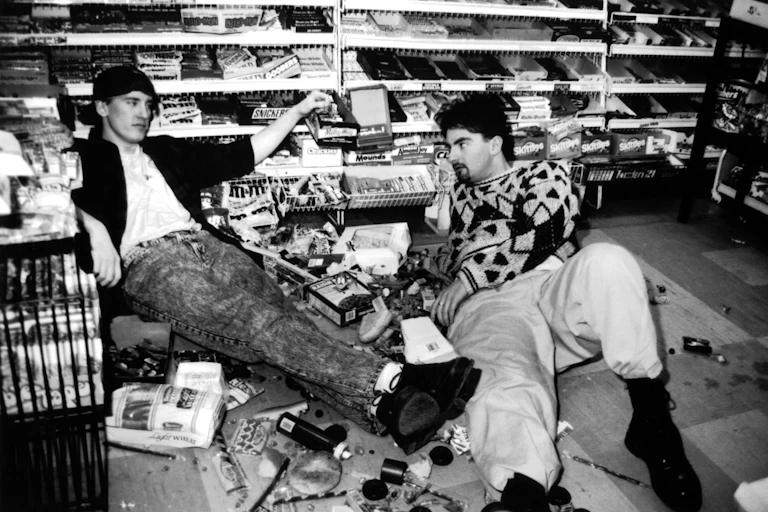Two men sit on the floor of a convenience store surrounded by spilled merchandise.