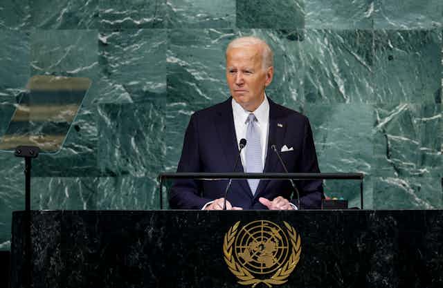 A white man in a blue suit stands at a lectern with the United Nations emblem on.