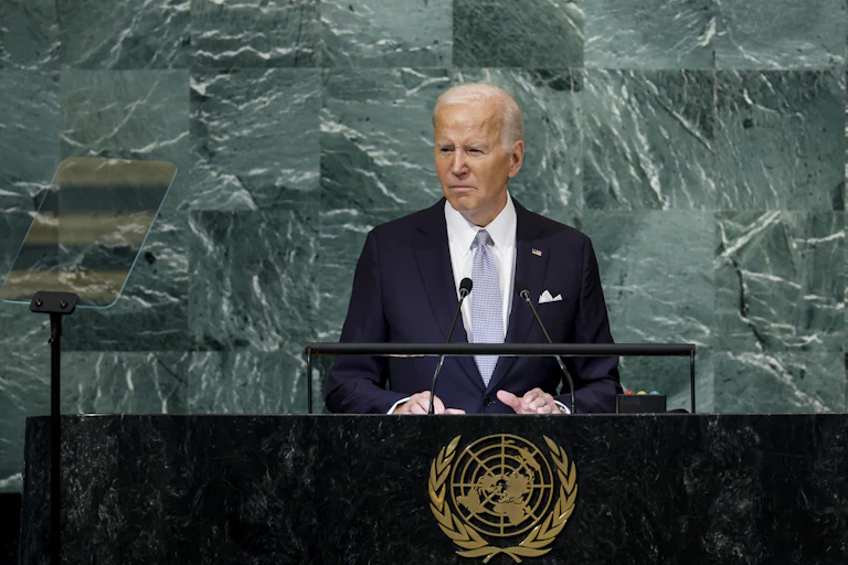 A white man in a blue suit stands at a lectern with the United Nations emblem on.