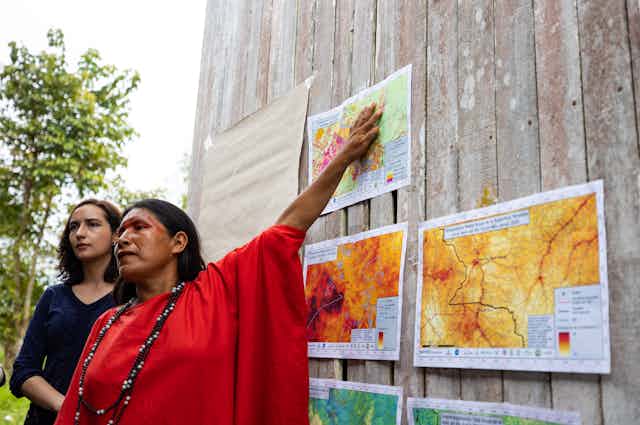 An Indigenous woman speaking to people outside points to maps posted on a wooded wall