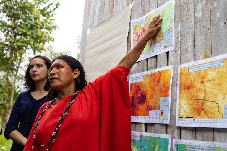 An Indigenous woman speaking to people outside points to maps posted on a wooded wall