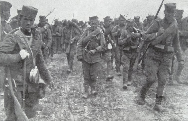 A black and white photo shows young looking men and boys in army uniform trudging through the mud.