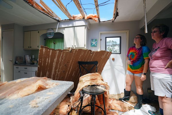 Две женщины стоят в поврежденной ветром кухне и смотрят на небо через недостающую часть крыши.