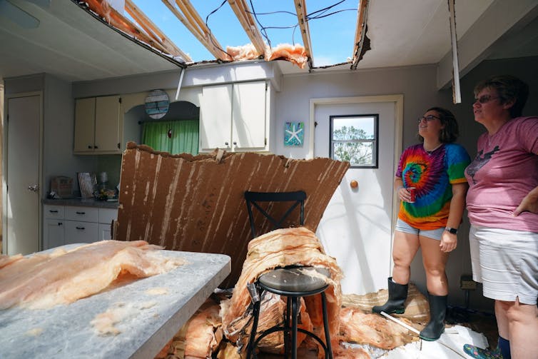 Two women stand in a wind-damaged kitchen looking up at the sky through a missing section of roof.