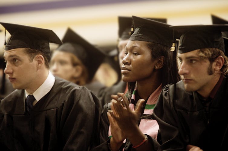 A Black woman wearing a black graduation cap and gown is seated in between two white male college graduates.