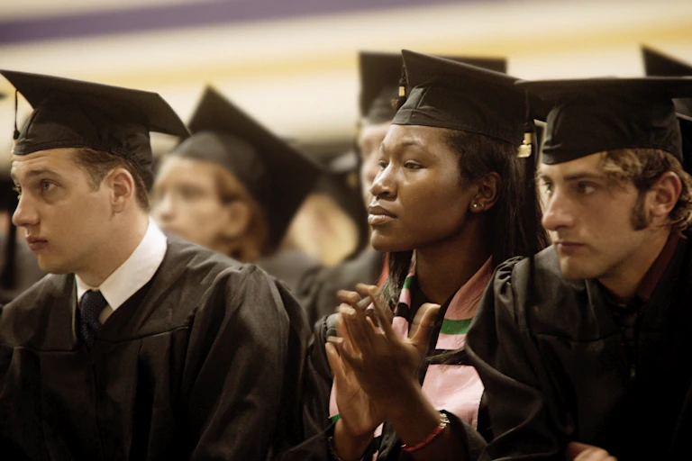 A Black woman wearing a black graduation cap and gown is seated in between two white male college graduates.
