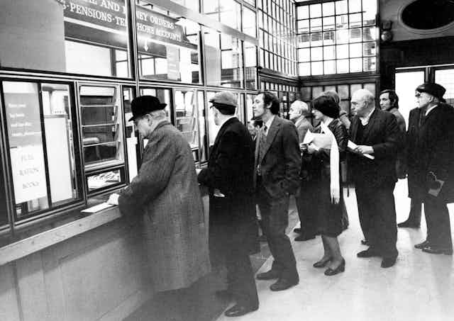 A queue of people waiting at a service booth in 1970s London.
