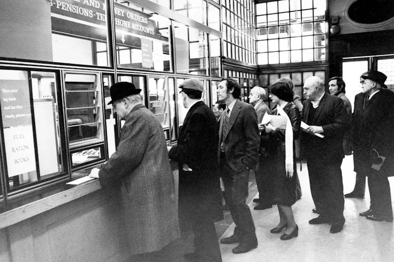 A queue of people waiting at a service booth in 1970s London.