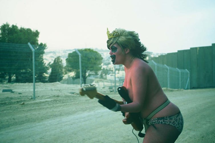 A woman in her underwear plays a guitar near a high security wall. She wears a wig and crown with exaggerated make-up.