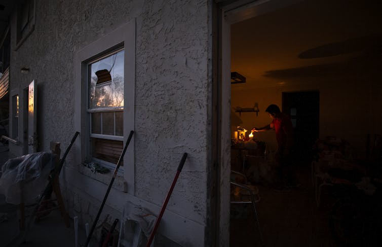 A person washes dishes by torchlight, with smashed windows visible outside.