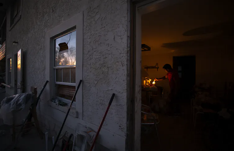 A person washes dishes by torchlight, with smashed windows visible outside.
