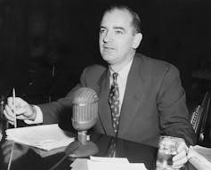A black and white photo shows a man in a suit and tie sat in front of a old-fashioned microphone.
