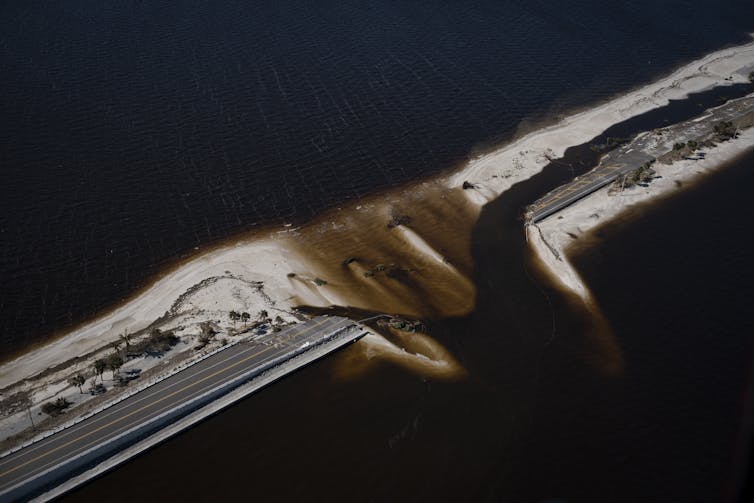 Aerial view of a causeway with water and sediment flowing through a gap in the center