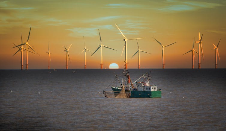 A fishing boat in front of an offshore wind farm against a deep sunset sky.