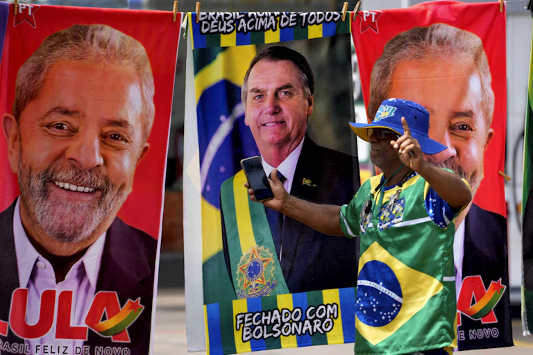 A man dressed in blue green and yellow stands in front of beach towels featuring the faces of two men.
