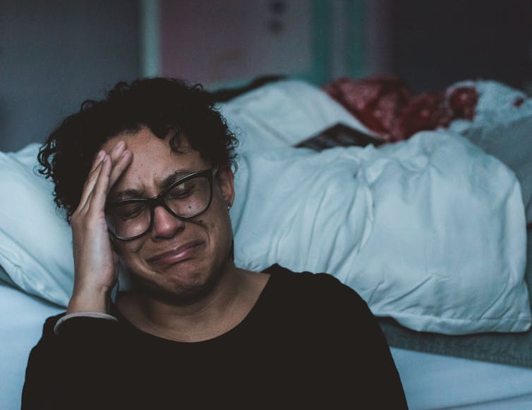 Brown-skinned person with hand on head crying, sitting against a bed.