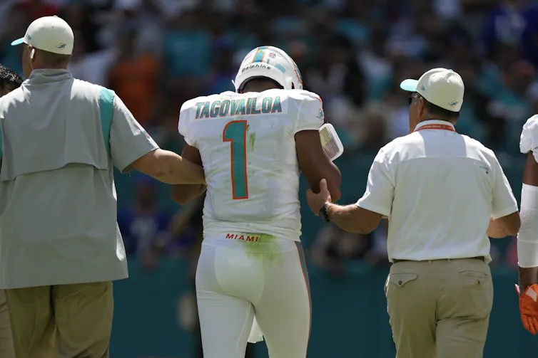 Two men in peaked caps assist a man in a white football uniform and helmet, seen from behind
