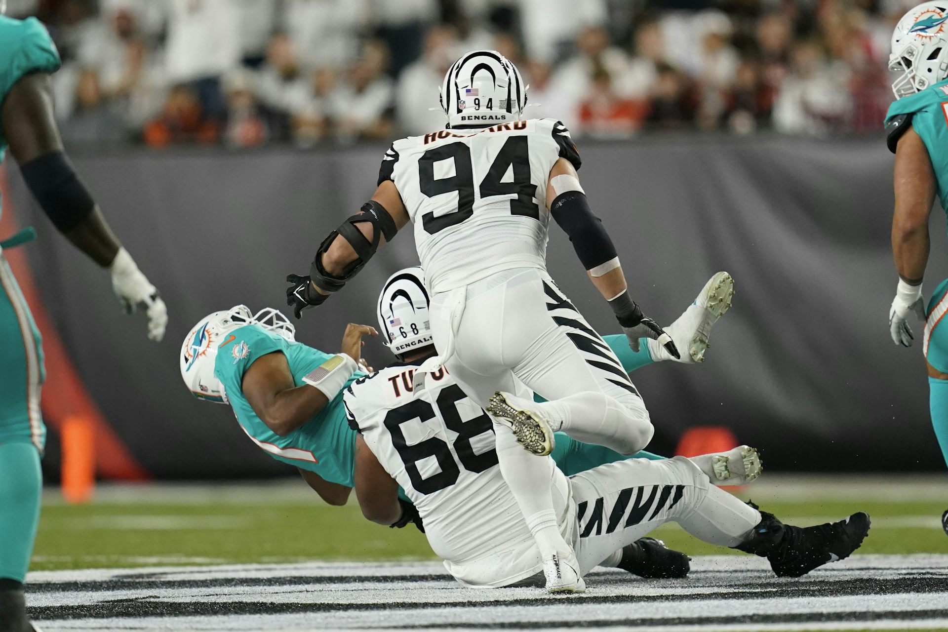 A football player in a teal uniform about to fall to the ground as a player in a white uniform tackles him, while another player in a white uniform runs towards them