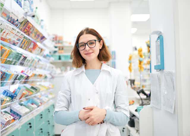 A young female pharmacist, wearing a white uniform and glasses, is standing inside a pharmacy, near shelves of over-the-counter drugs. 
