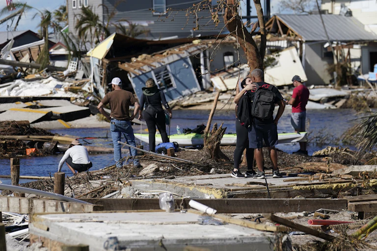 People stand on a damaged dock, observing wreckage all around them.