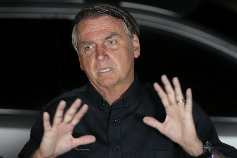 A man in a black suit holds up his hands as he talks.