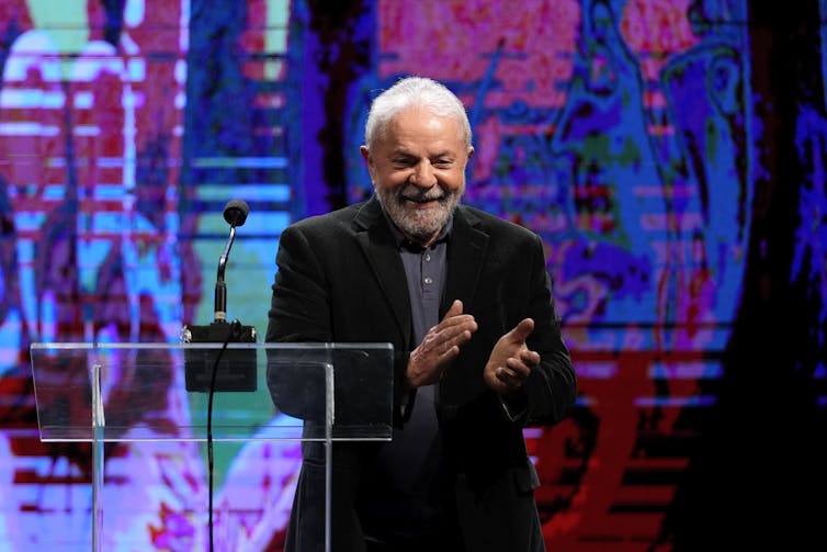 A grey haired man with a beard smiles and claps from behind a podium.