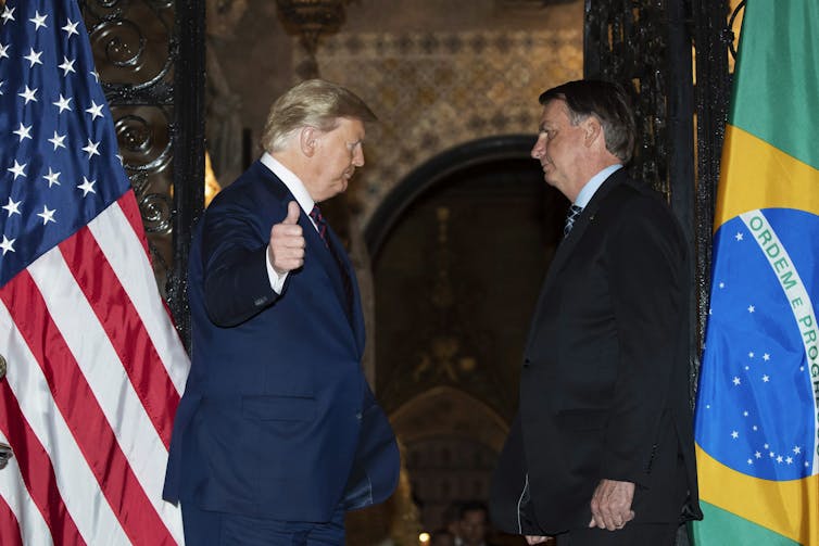 A blond man gives the thumbs up while standing next to a dark-haired man. The U.S. and Brazilian flags are behind them.