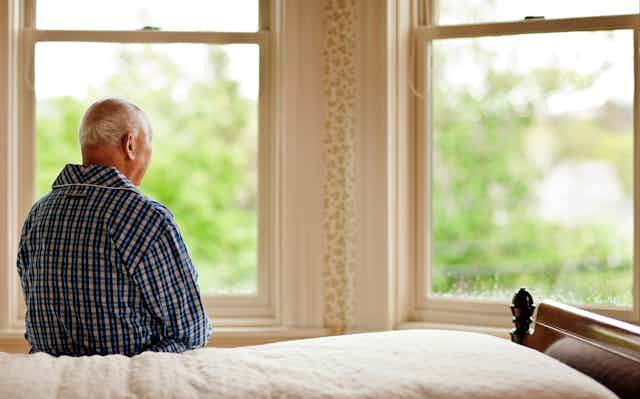 An elderly man sits on a bed facing away from the camera. He is looking out of a window. 