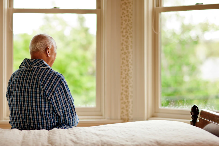 An elderly man sits on a bed facing away from the camera. He is looking out of a window.