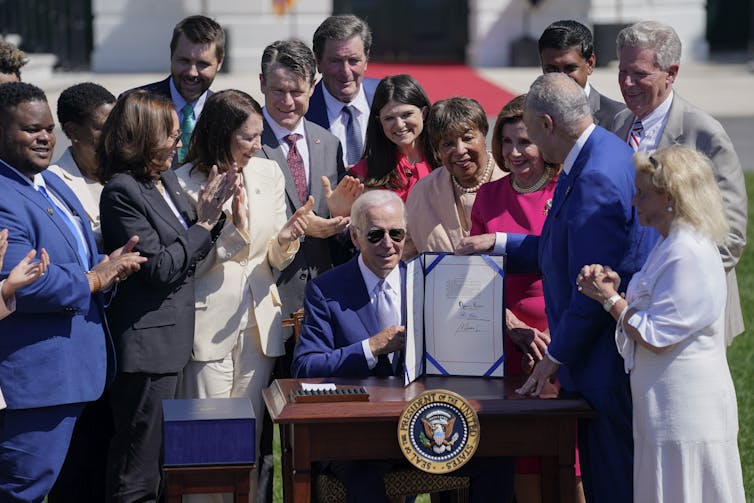 A white-haired man in a suit and sunglasses sits at a desk surrounded by people, with a document standing on the desk.