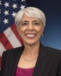 A white-haired woman smiles in a formal portrait with an American flag in the background.
