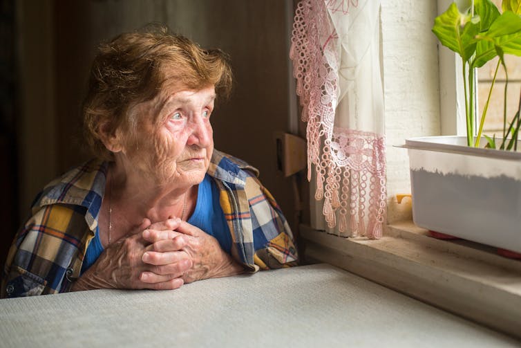 An elderly woman sits at a table looking out of a window. There is a green plant on the windowsill.