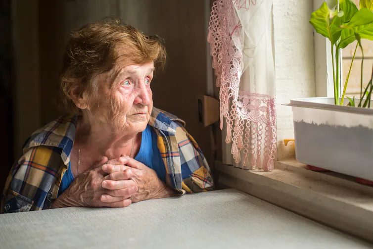 An elderly woman sits at a table looking out of a window. There is a green plant on the windowsill.