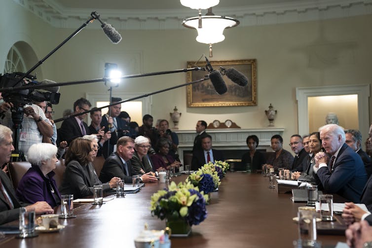 People in business attire sit around a long conference table with microphones placed above them.