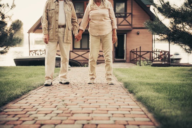 An elderly couple stand in front of a house holding hands.