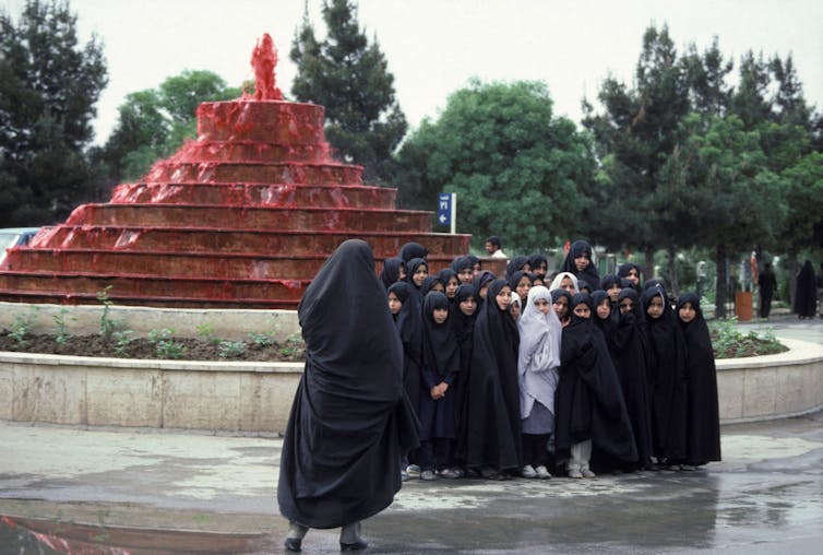 A group of girls in black robes poses in front of a fountain as a woman takes their photograph.