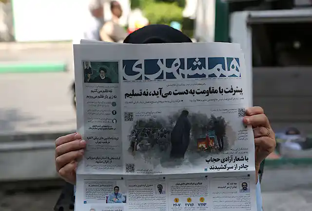 A woman holds a newspaper close to her face in Iran. Her face is not visible, just her hands and the top of her head.