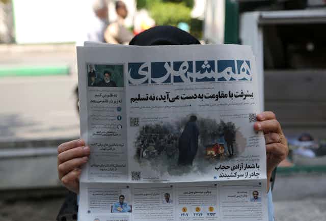 A woman holds a newspaper close to her face in Iran. Her face is not visible, just her hands and the top of her head.