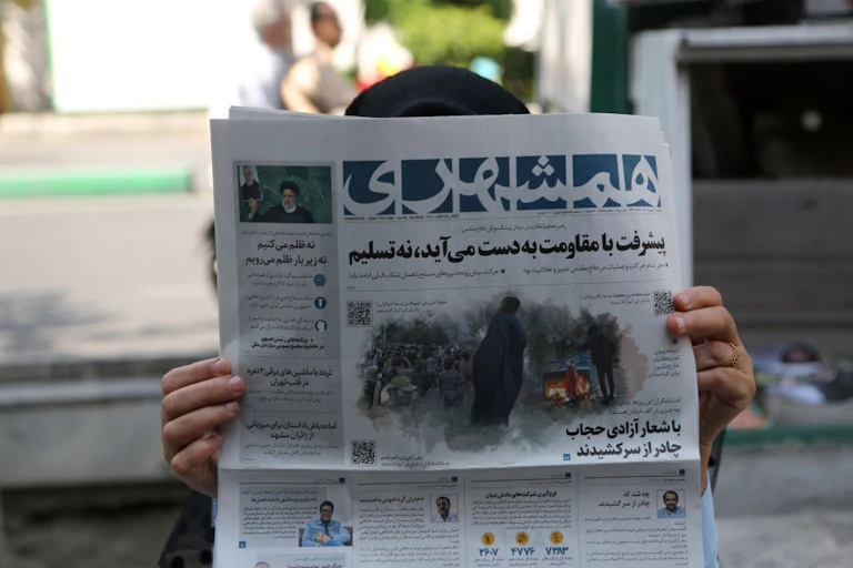 A woman holds a newspaper close to her face in Iran. Her face is not visible, just her hands and the top of her head.