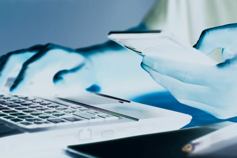 A person typing on laptop with smartphone in the other hand