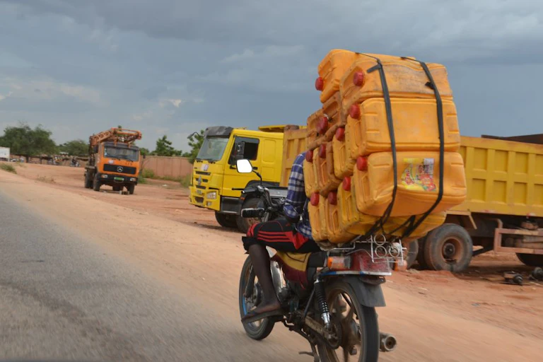 A motorcyclist riding past parked lorries with jerrycans of fuel strapped to the back of his motorbike.
