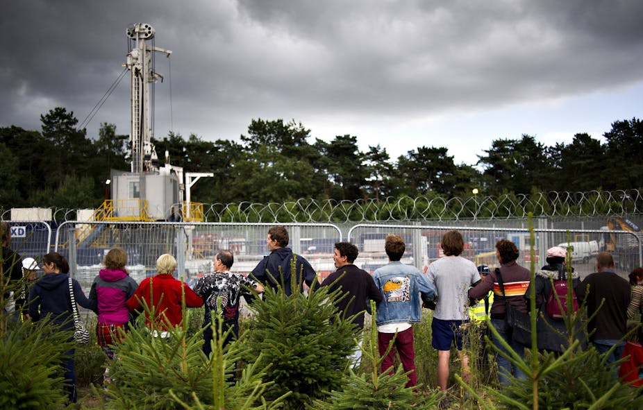 Protestors standing in a line outside the perimeter of a fracking site against a forested backdrop.