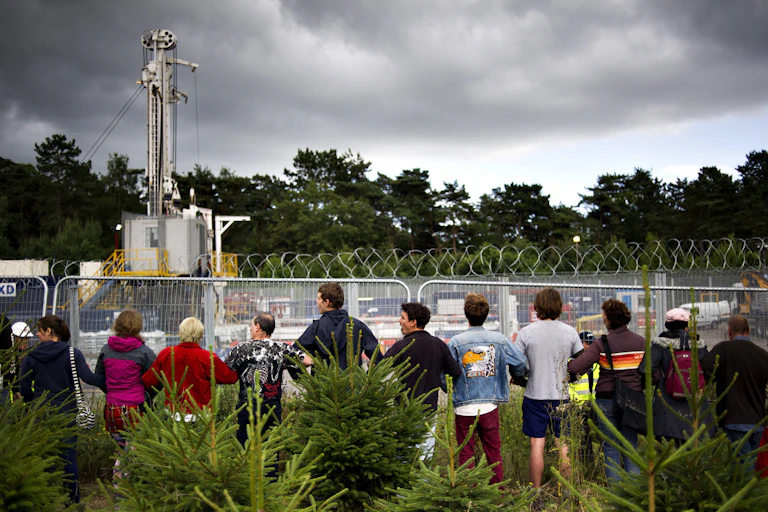 Protestors standing in a line outside the perimeter of a fracking site against a forested backdrop.